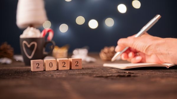 Low angle view of 2022 sign on wooden cubes with christmas holiday decorations in background and female hand writing new year plans in a notebook.