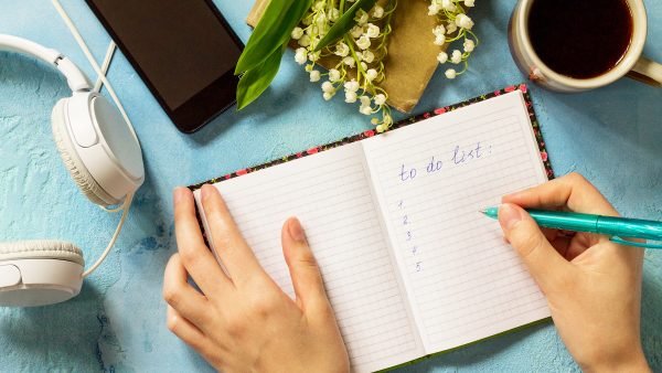 Smartphone in female hands and writing in notebook, lilies of the valley and headphones on stone table. Top view flat lay background with copy space.