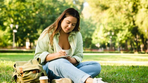 Imagination concept. Young freelancer woman student doing homework, e-learning, preparing for test exam, writing diary poems novels relaxing on college university lawn park